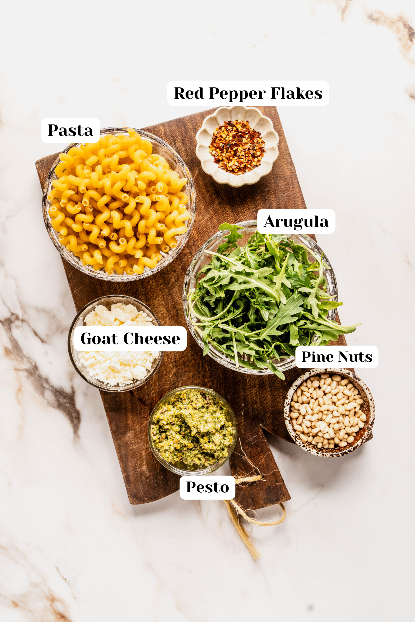 Overhead shot of ingredients for pesto goat cheese pasta arranged on a wooden cutting board over a white marble background, including a bowl of cavatappi pasta, fresh arugula, crumbled goat cheese, pine nuts, red pepper flakes, and a jar of vibrant green pesto.