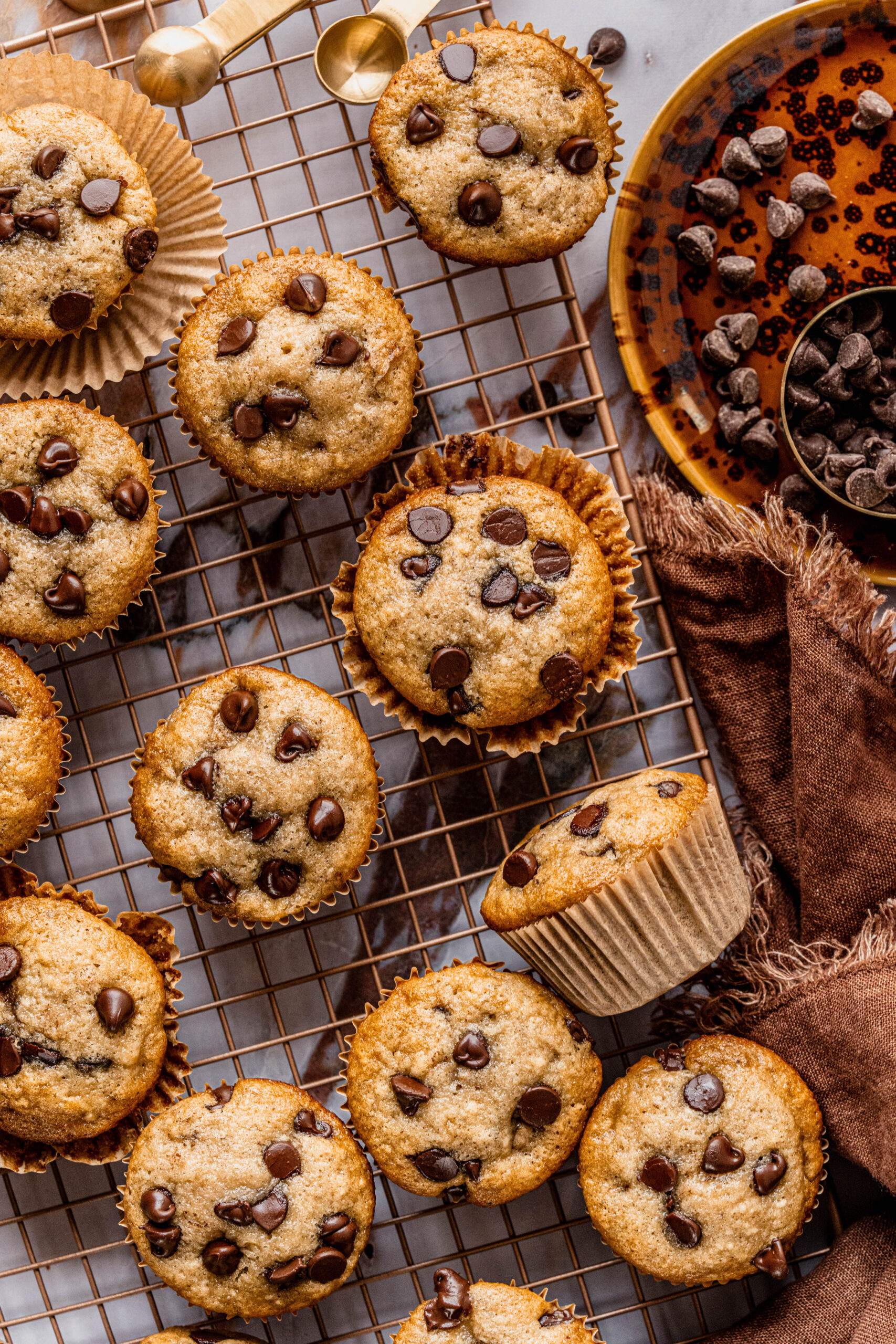 Overhead view of banana chocolate chip gluten free muffins arranged on a cooling rack, with golden tops studded with chocolate chips, surrounded by a bowl of chocolate chips and a brown linen napkin on a marble surface.