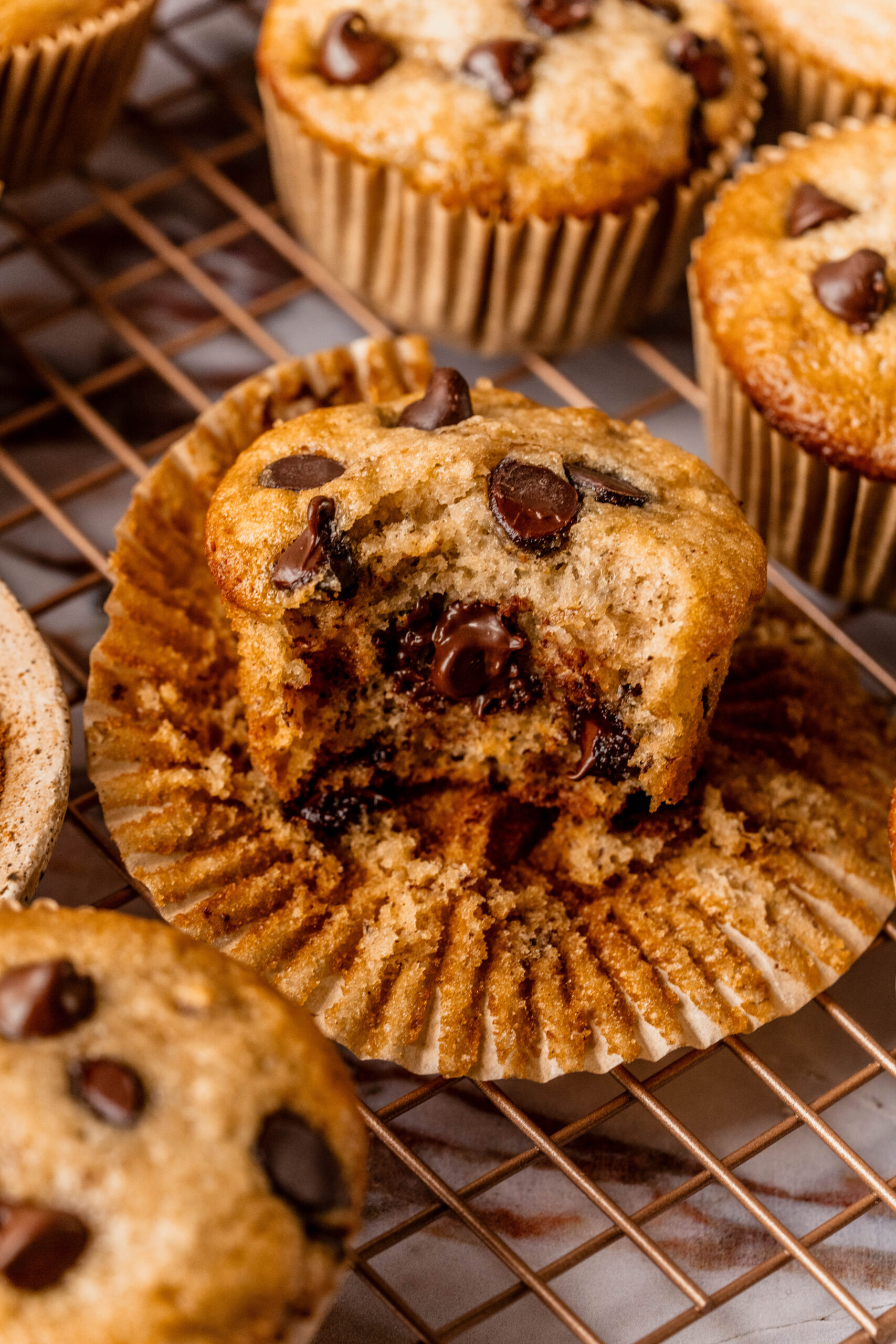 Overhead close-up of banana chocolate chip gluten free muffins on a cooling rack, with one muffin broken open to reveal a soft, moist crumb and melty chocolate chips inside, styled on a marble background.