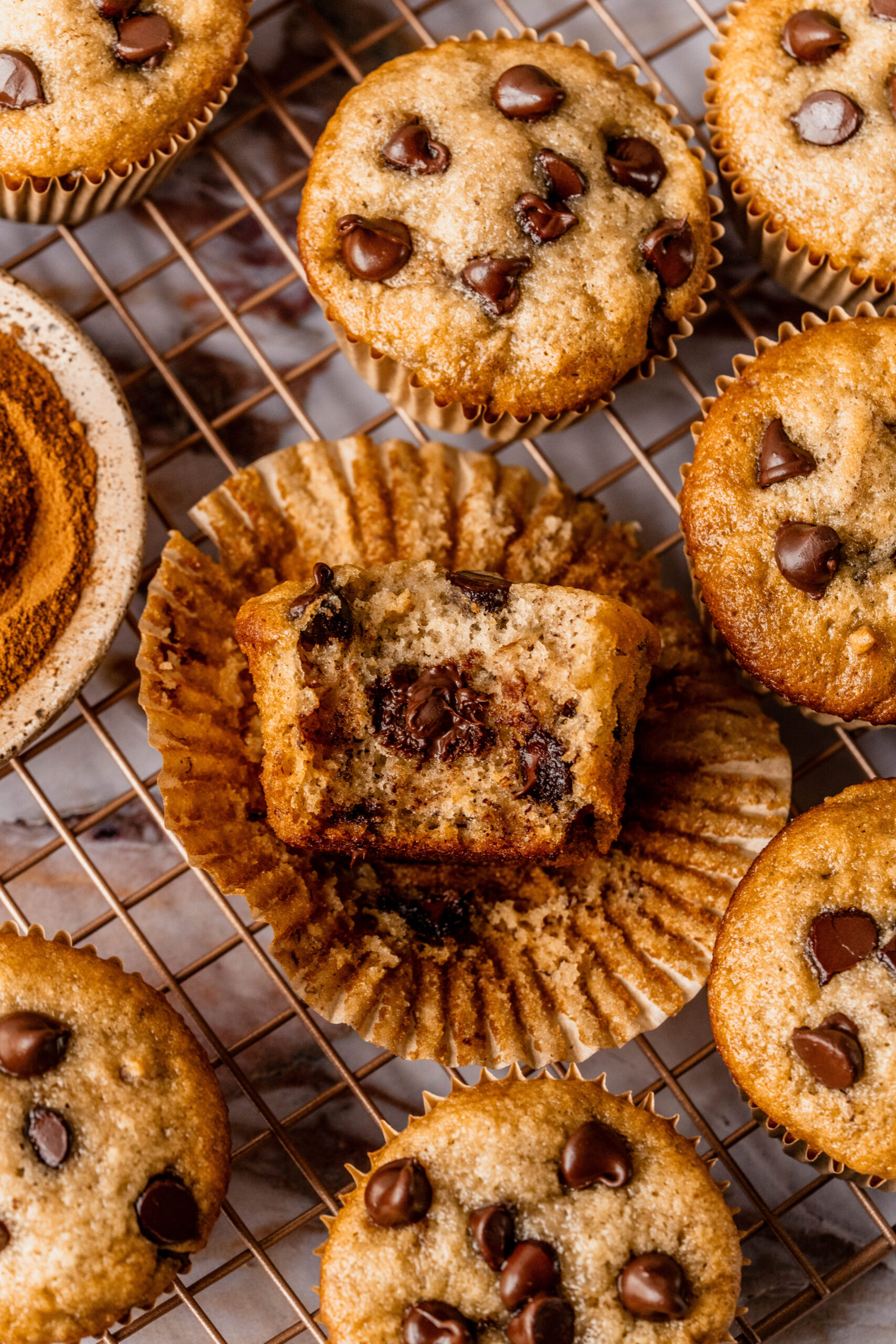 Close-up overhead of banana chocolate chip gluten free muffins on a cooling rack, with one muffin split open to show a soft, moist crumb filled with melted chocolate chips, styled on a marble background.
