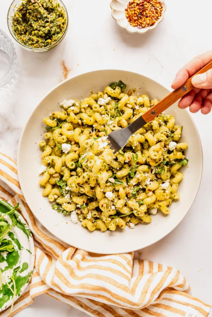 Bright overhead shot of pesto goat cheese pasta made with cavatappi, tossed in green pesto and arugula, topped with crumbled goat cheese, toasted pine nuts, and red pepper flakes, with a hand holding a fork lifting a bite from a bowl on a light marble surface.