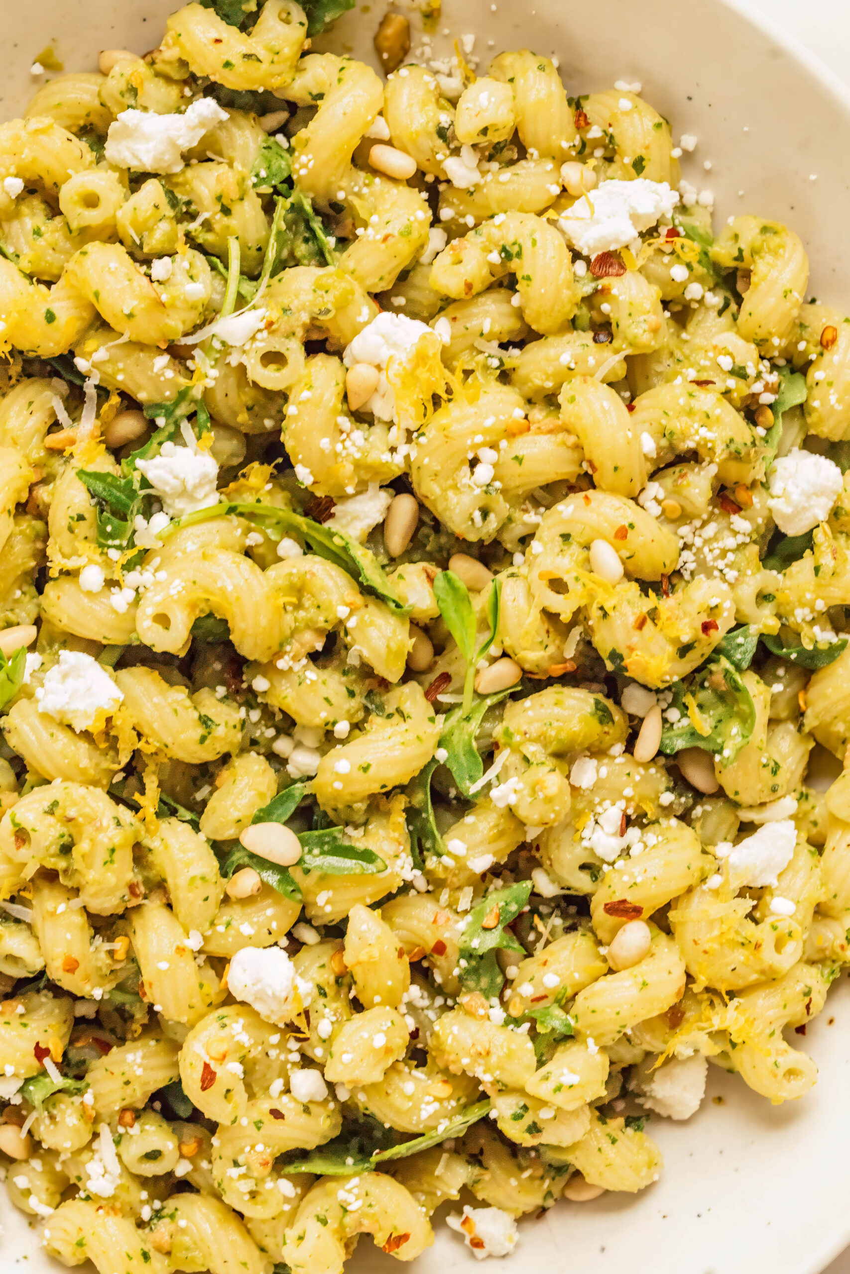 Macro close-up of pesto goat cheese pasta with cavatappi coated in vibrant pesto, tossed with fresh arugula, and topped with crumbled goat cheese, toasted pine nuts, red pepper flakes, and bright lemon zest.